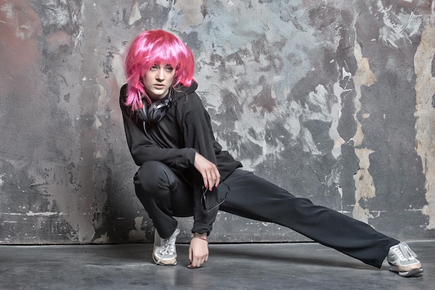Fitness, activity concept. girl in sport suit stretch leg on abstract wall. fashion, beauty, look. woman wear pink wig hair with headphones. music, technology, entertainment concept.