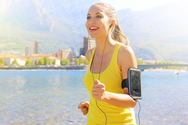 Fitness athlete woman running and wearing phone armband with touchscreen during cardio workout on lake.