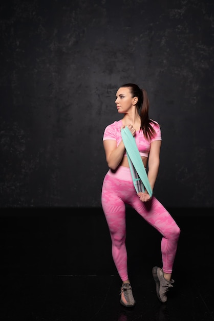 Fitness athletic girl. Woman in fashionable sportswear, standing against a black background, after a workout. A girl with a strong muscular body, enjoys her body