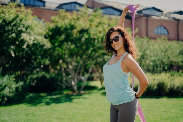 La donna atletica di forma fisica fa allenamento con l'elastico vestito con occhiali da sole alla moda di abbigliamento sportivo pone all'esterno