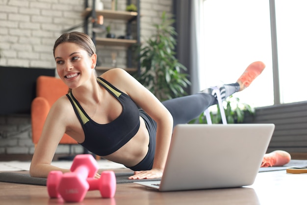 Fitness beautiful slim woman doing side plank with resistance\
band and watching online tutorials on laptop, training in living\
room. stay at home activities.