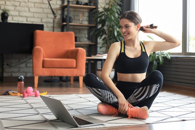 Foto fitness bella donna magra è seduta sul pavimento con manubri e bottiglia d'acqua utilizzando il computer portatile a casa nel soggiorno. restare a casa attività.