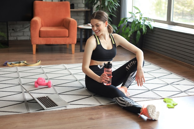 Fitness beautiful slim woman is sitting on the floor with dumbbells and bottle of water using laptop at home in the living room. Stay at home activities.