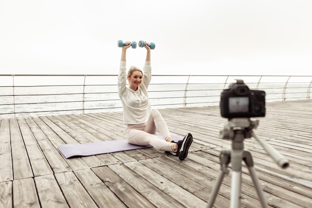 Fitness bloggen. Jonge gezonde sportvrouw die training op camera met statief opneemt en laat zien hoe ze op het strand moet oefenen. vloggen