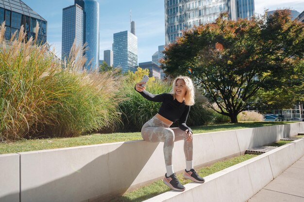 Fitness blonde lady making selfie using mobile outdoors Athlete woman in sportswear relaxing after workout at the street with skycrapers on backgorund