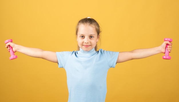 Fitness child portrait of sporty little girl with dumbbells over yellow studio isolated background g...