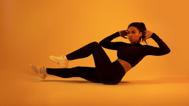 Фитнес-концепция Подтянутая чернокожая женщина делает хруст живота локтями на коленях, тренируясь на полу на неоновом оранжевом фоне
