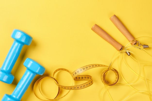 Fitness concept with blue dumbbells, measuring tape and jump rope on a yellow background