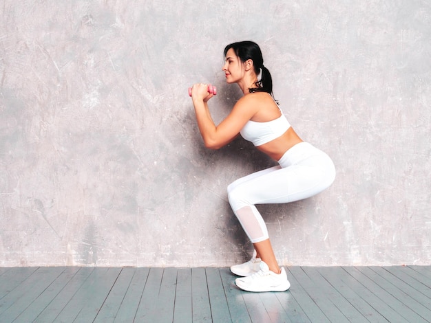 Fitness confident woman in white sports clothing Sexy young beautiful model with perfect body Female posing near grey wall in studio Stretching out before training Making squats with dumbbells