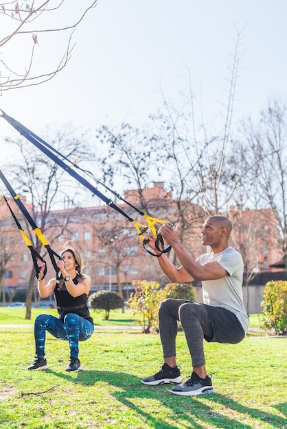 公園でtrxフィットネスストラップを使って脚の運動をしているフィットネスカップル。屋外で運動する多民族の人々。