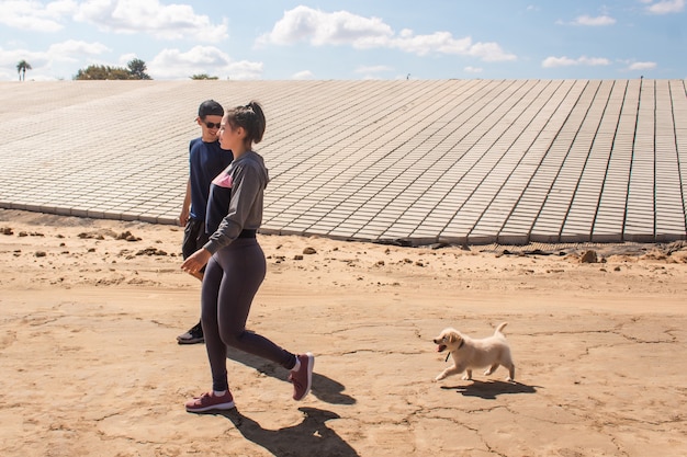 Fitness couple walking in the sand while their puppy follows them from behind.