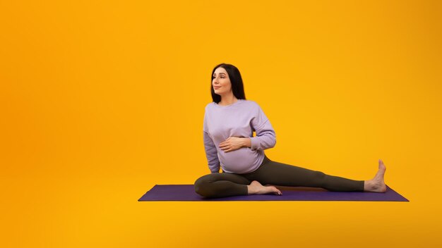 Fitness during pregnancy pregnant lady sitting on yoga mat and stretching legs looking aside at free space panorama