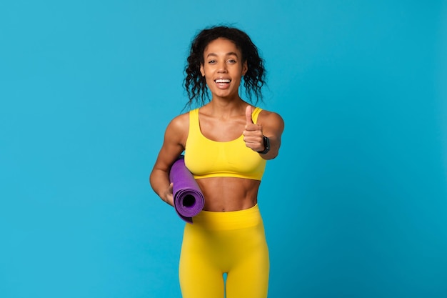 Fitness enthusiast black lady with yoga mat on blue background