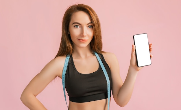 Fitness girl smiling and holding a phone with a mockup and posing hold measure tape in black sportswear on a pink surface