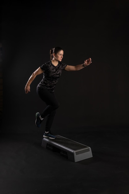 Photo fitness girl on a step platform pulls her leg go in for sports on a black background joyful