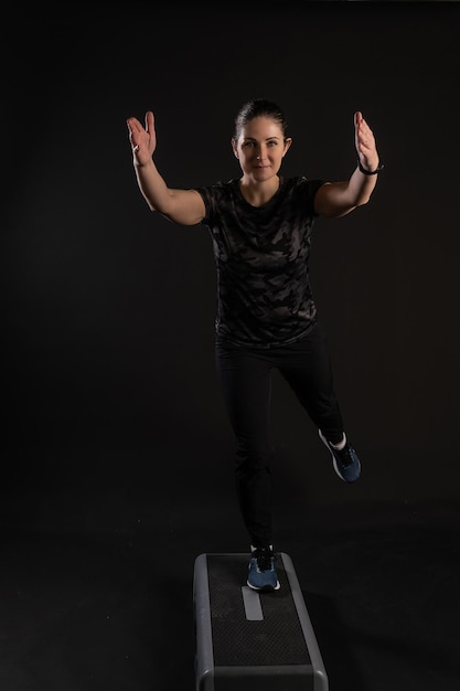 Fitness girl on a step platform pulls her leg Going in for sports on a black background Happy