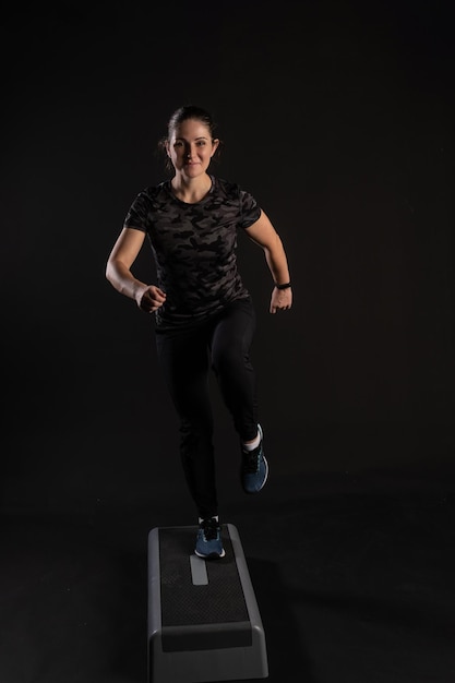 Fitness girl on a step platform stretches her arms Go in for sports on a black background with