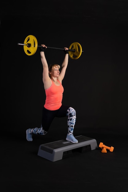 Photo fitness girl on a step platform with a barbell with dumbbells go in for sports on black background