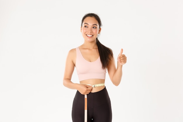 Fitness, healthy lifestyle and wellbeing concept. Portrait of satisfied smiling, cute asian girl in sportswear, showing thumbs-up after measuring waist with tape measure, lost weight.