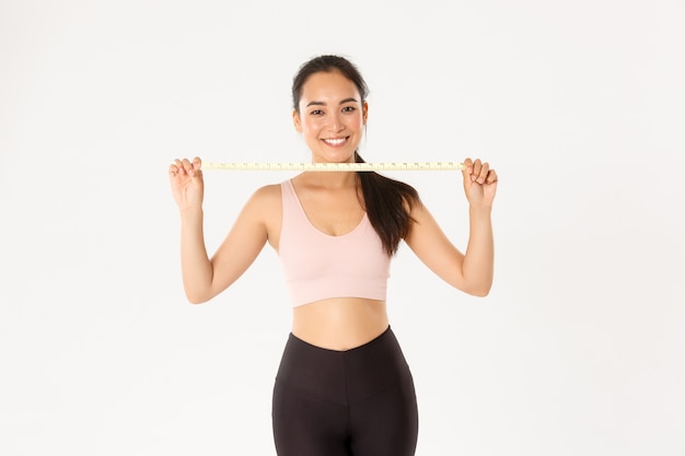 Fitness, healthy lifestyle and wellbeing concept. Portrait of smiling asian brunette sports girl, female athelte in activewear showing measuring tape, losing weight with exercises 