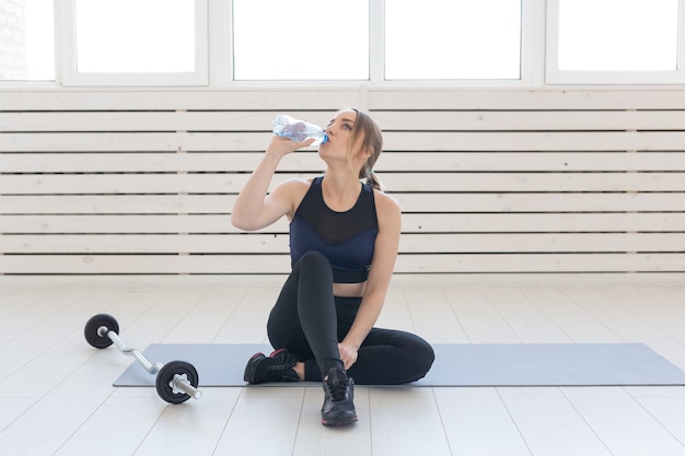 Fitness, healthy and sport concept - young woman sitting cross-legged and drinking water from bottle after workout.