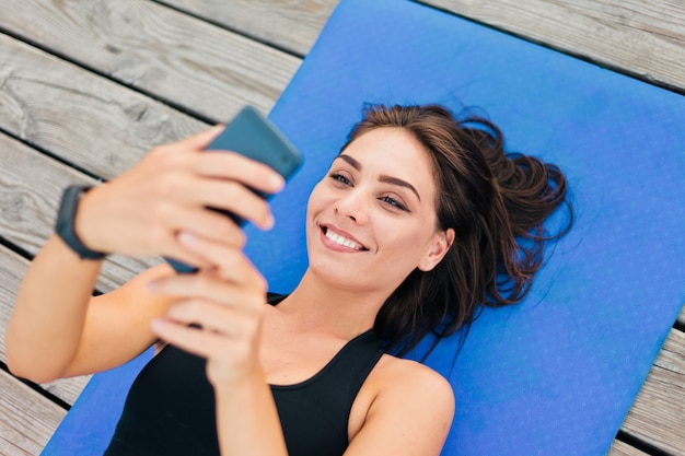 Photo fitness healthy woman in sportswear is lying on the mat and holding a smartphone in her hands on wooden boards.