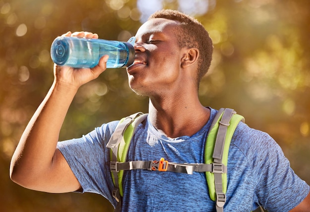 Fitness hiking and black man with water bottle for hydration while outdoor in nature with backpack for travel trekking and adventure Thristy male in a forest for a hike workout and training