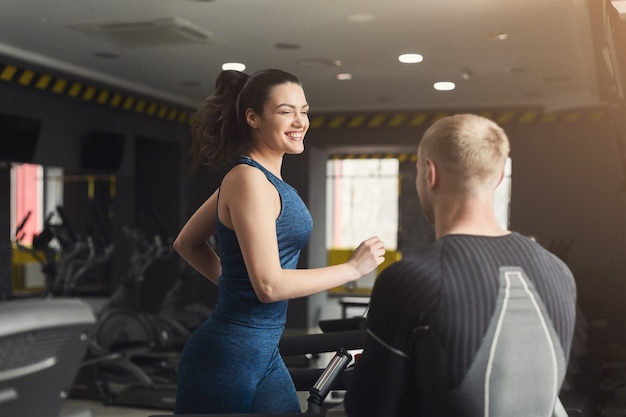 Fitness instructor helps young woman on elliptical trainer. Coach discussing workout plan with girl on treadmill. Cardio workout in gym, healthy lifestyle, copy space