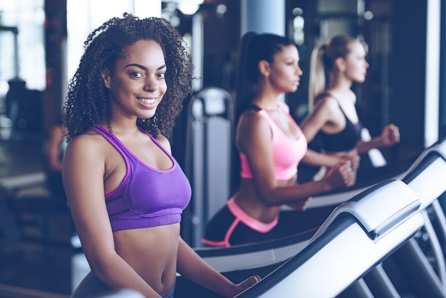 fitness is her lifestyle. beautiful young cheerful african woman in sportswear looking at camera with smile while running on treadmill at gym with other women