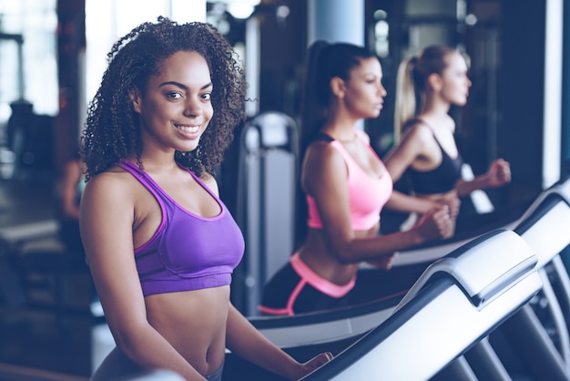 Fitness is her lifestyle. Beautiful young cheerful African woman in sportswear looking at camera with smile while running on treadmill at gym with other women