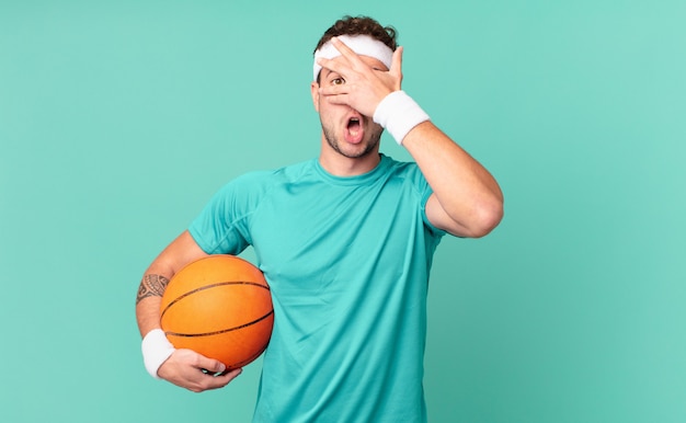 Fitness man looking shocked, scared or terrified, covering face with hand and peeking between fingers