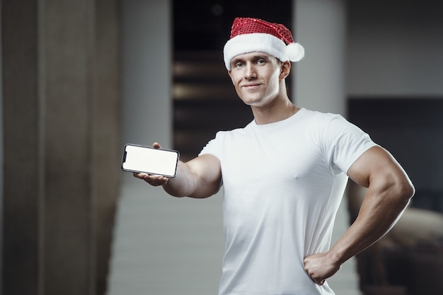 Fitness man in Santa Claus hat costume in gym with cell phone.