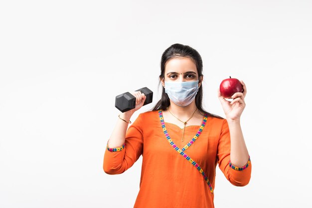 유행병 개념의 피트니스 - 예쁜 인도 소녀는 덤벨과 함께 운동하고 사과를 보여주는 동안 의료용 안면 마스크를 착용합니다