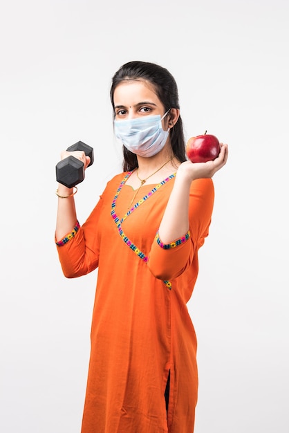 유행병 개념의 피트니스 - 예쁜 인도 소녀는 덤벨과 함께 운동하고 사과를 보여주는 동안 의료용 안면 마스크를 착용합니다