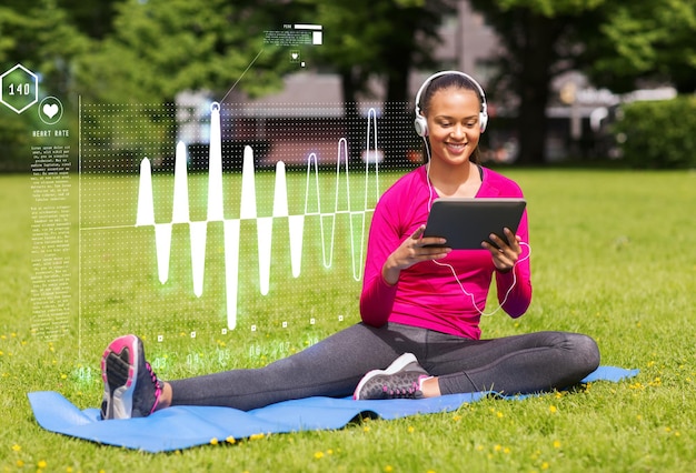 fitness, park, technologie en sport concept - lachende Afro-Amerikaanse vrouw met tablet pc-computer en koptelefoon op mat buitenshuis