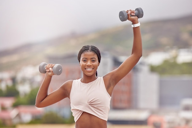 ボディービルと強力な腕のためのダンベルで重量挙げをしているフィットネスの肖像画と女性顔の正面とアフリカ系アメリカ人の女性のボディービルダーが、都市での強さのために運動またはトレーニングを行っています