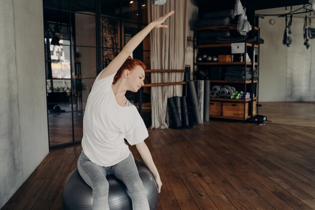 Fitness practice. Young active red haired woman in sportswear sitting on large silver exercise fitball and working out in sports club or studio, doing stretching exercises by lifting up one hand