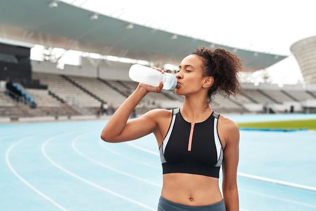 フィットネス ランニングとスタジアムでボトルから飲むトラック上の女性は、マラソン トレーニングや運動中にリラックスします。トレーニング タイムの健康とウェルネス。水のあるエリアで健康な女性スポーツ ランナー