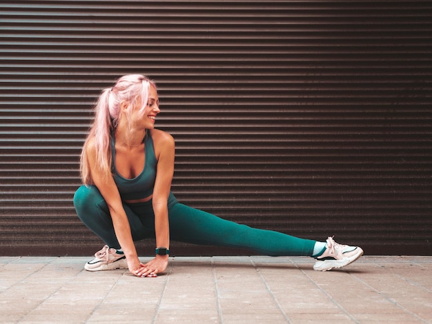 Fitness smiling woman in green sports clothing with pink hair Young beautiful model with perfect bodyFemale posing in the street near roller shutter wall Stretching out before training