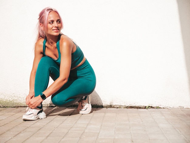 Fitness smiling woman in green sports clothing with pink hair Young beautiful model with perfect bodyFemale posing in the street near white walltying shoelace on running shoes before training