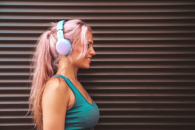 Fitness smiling woman in green sports clothing with pink hair Young beautiful model with perfect bodyFemale in the street near roller shutter wallListening music in wireless headphones