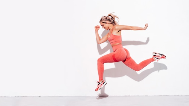 Fitness smiling woman in pink sports clothing Young beautiful model with perfect body Female posing in the street near white wall at summer sunny day Cheerful and happy Jumping and running