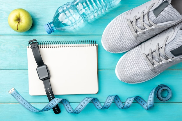 Fitness sneakers, notebook, green apple, tape measure and bottle of water on blue wooden background