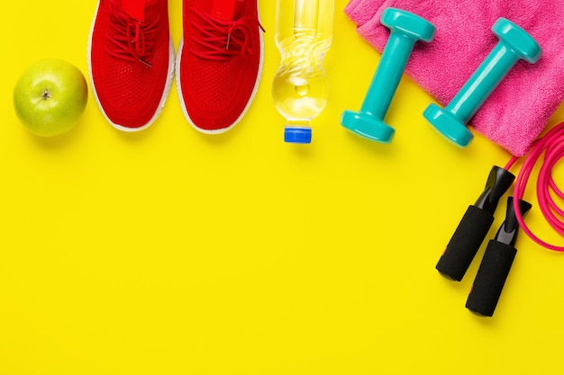 Fitness and sport concept jump rope green apple dumbbells bottle of water red sneakers and pink towel on bright yellow background free space