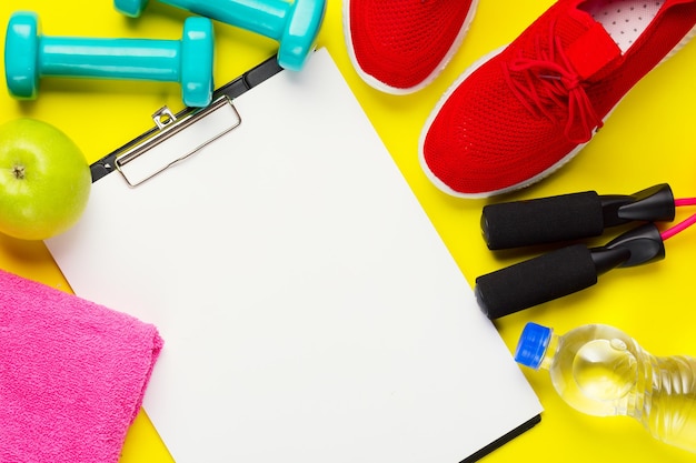 Fitness and sport concept. Red sneakers, apples, jump rope, dumbbells, bottle of water, pink towel and sheet of blank paper on yellow background.