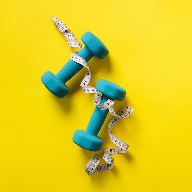 Fitness, sport concept with dumbbells and measuring tape over yellow background