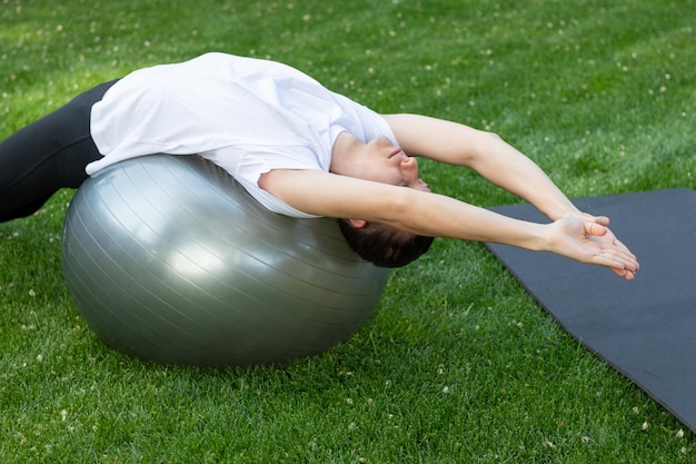 Fitness sport en gezonde levensstijl concept Jonge man die zijn rug strekt op een balansbal in de tuin
