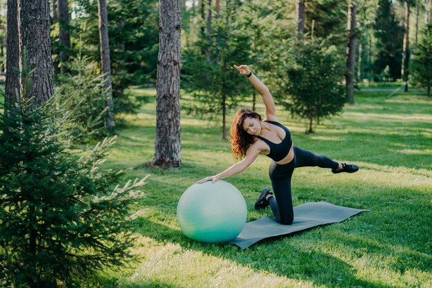 Fitness sport en gezonde levensstijl concept Outdoor shot van slanke brunette Europese vrouw doet pilates en balans oefening met fit bal kantelt opzij poses in groen bos op gras op karemat