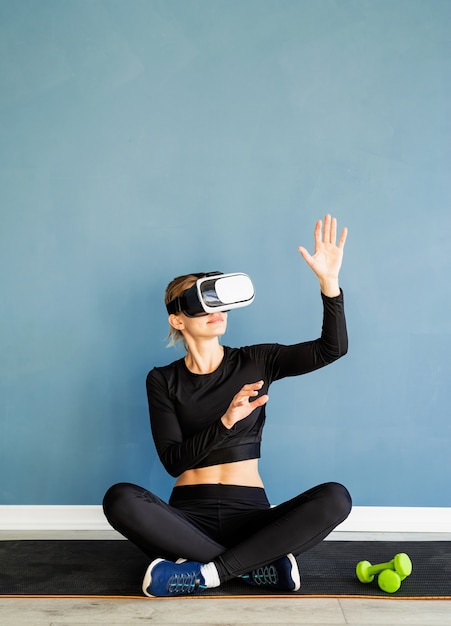 Fitness, sport en technologie. Jonge atletische vrouw VR bril zittend op fitness mat met behulp van het interactieve menu Vr bij blauwe achtergrond