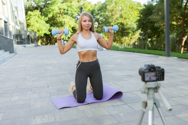 Fitness, sport en video bloggen concept. Vrouwelijke sportblogger met camera op statief die outdoor fitnesstraining met halters opneemt